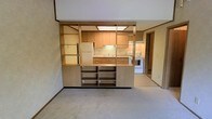 This single-floor space showcases an open-concept layout that integrates the living area and kitchen into one cohesive environment. A defining architectural feature is the freestanding built-in unit that serves as a partial divider between the two spaces. This unit combines open shelving on both sides with integrated cabinetry, offering substantial storage while maintaining visual connectivity throughout the space.

The kitchen is equipped with light wood cabinetry, a white refrigerator, a white range, and a sink with under-cabinet lighting that illuminates the workspace. Upper cabinets line the walls, providing ample storage, while the layout is practical and efficiently arranged.

The living area features neutral wall-to-wall carpeting in a light beige tone, contributing to a warm and understated ambiance. The walls are finished in a textured neutral tone, and overhead fluorescent lighting panels provide consistent illumination. A doorway visible toward the rear of the kitchen leads to an additional room or hallway, suggesting further usable space beyond what is immediately visible. Overall, the space has a functional, straightforward design with a cohesive neutral color palette throughout.