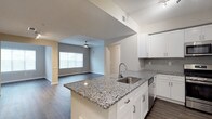 This single-floor residence offers a thoughtfully designed open-concept layout that seamlessly connects the kitchen and living areas. The kitchen is a standout feature, boasting granite countertops with a striking salt-and-pepper finish, a stainless steel undermount sink set within a central peninsula, and crisp white cabinetry with brushed nickel hardware. A classic subway tile backsplash in a soft gray tone adds a refined touch, while built-in appliances contribute to the kitchen's clean, modern aesthetic. The peninsula doubles as a casual dining or prep space, making it both functional and stylish. Dark wood-style flooring flows throughout the open living area, providing a warm contrast to the light-toned walls. The living space is generously proportioned and benefits from natural light streaming in through a large window fitted with horizontal blinds. A ceiling fan is centrally positioned overhead, enhancing comfort and airflow. The textured ceiling adds a subtle architectural detail consistent throughout the space. Neutral wall tones and clean sightlines give the interior a fresh, move-in-ready feel that is both inviting and practical.