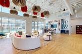 Located at 9910 W Montebello Ave in Phoenix, AZ, this single-story community clubhouse is a thoughtfully designed social hub that balances modern aesthetics with functional versatility. The space is anchored by soaring ceilings with an exposed structural grid, from which a striking collection of sculptural pendant lights in neutral tan and bold red tones cascade dramatically overhead, serving as a defining architectural focal point.

The main lounge area features warm herringbone-patterned hardwood flooring and is furnished with a curved white sectional sofa, accent chairs, and a patterned area rug, creating an inviting and stylish gathering zone. Floor-to-ceiling glass walls and sliding doors along one side flood the interior with abundant natural light while offering direct views of the outdoor pool and courtyard area.

A dining and bar zone includes a tall communal table with bar-height seating, positioned adjacent to a built-in feature wall of white open shelving accented by geometric blue tile, housing a large mounted television and a beverage refrigerator below.

A private conference or meeting room, enclosed by sliding frosted-glass and terracotta-toned panels, offers an intimate space with sage green walls, herringbone flooring, globe pendant lighting, and a white conference table with upholstered seating. The entryway features decorative etched-glass partitions and graphic wall art, setting a vibrant, contemporary tone upon arrival.