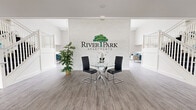 Located at 1616 W Bender Rd in Glendale, Wisconsin, this expansive single-floor community clubhouse and leasing center for River Park Apartments offers a generously proportioned, open-plan interior designed to impress. The space is defined by soaring double-height ceilings that create an airy, grand atmosphere throughout.

The entry foyer features a striking textured accent wall prominently displaying the River Park Apartments branding, complemented by wood-look flooring in a neutral gray tone that flows seamlessly throughout the entire space. White-railed staircases flank the central area, adding architectural symmetry and elegance.

A central lounge area is anchored by a floor-to-ceiling white fireplace, surrounded by plush seating in soft blue and lavender tones with gold-accented furnishings. Adjacent to this, a sunlit gathering area benefits from a dramatic wall of expansive floor-to-ceiling windows, flooding the interior with natural light and offering views of the surrounding property.

Bold cobalt blue accent walls serve as a backdrop for large-scale floral artwork, adding a vibrant, contemporary character. The leasing area near the entrance features a round glass table with modern chrome chairs. Throughout, the design balances clean white architectural elements with carefully curated furnishings, creating a welcoming and polished community space.