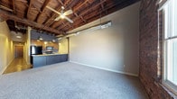 This single-floor loft-style space showcases a striking open layout that combines industrial character with modern design sensibilities. The most commanding feature is the exposed wood beam ceiling, which adds warmth and architectural depth to the interior. A ceiling fan is centrally mounted, providing both function and a rustic focal point overhead. Visible ductwork runs along the ceiling, reinforcing the industrial aesthetic that defines the space.

The main living area is finished with plush, neutral-toned carpet that softens the rawness of the industrial elements and creates a comfortable, inviting atmosphere. The open floor plan allows for flexible furniture arrangements and an unobstructed flow throughout the space.

The kitchen is positioned at one end of the open layout and features dark cabinetry paired with modern black appliances, including a refrigerator, microwave, and stove. Pendant lighting hangs above the kitchen area, providing focused illumination and adding a stylish touch. A kitchen counter extends to create a natural transition between the cooking area and the living space.

Neutral gray walls complement the warm wood tones of the ceiling, creating a balanced and cohesive palette throughout. The combination of exposed structural elements, open planning, and contemporary finishes gives this space a distinctive urban loft character that is both functional and visually compelling.