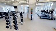 This modern fitness center is designed to support a comprehensive workout experience within a bright, open layout. The space features large floor-to-ceiling windows along one wall, flooding the interior with natural light and offering views of the surrounding cityscape. The clean white walls and light-colored rubberized flooring create a fresh, professional atmosphere throughout.

The facility is well-stocked with a full range of free weights, including a neatly organized dumbbell rack accommodating various weight increments, ideal for strength training exercises. A colorful medicine ball tower stands prominently, offering a variety of weighted balls for functional fitness and conditioning workouts. Along the windowed wall, a row of treadmills is lined up to provide ample cardio options while enjoying the natural light.

Exposed ceiling pipes and track lighting give the space an industrial-modern aesthetic that complements its contemporary fitness equipment. Wall-mounted flat-screen monitors are positioned for easy viewing during workouts. Additional equipment, including cable machines, can be spotted in the background, suggesting a diverse range of training options. Overall, this fitness center combines functionality with a clean, inviting design to accommodate a wide variety of exercise routines.