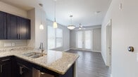 This single-floor residence showcases a thoughtfully designed open-concept layout where the kitchen flows seamlessly into the living and dining areas. The kitchen is well-appointed with dark espresso cabinetry, granite countertops, a stainless steel appliance suite including a refrigerator, range, microwave, and beverage cooler, complemented by a tile backsplash and pendant lighting over the central island with an integrated sink. Built-in shelving near the kitchen adds practical display and storage space.

The living area features wood-style plank flooring, a ceiling fan, and French doors that invite abundant natural light while providing outdoor access. Multiple windows fitted with horizontal blinds further brighten the space, contributing to an open and airy ambiance throughout.

The bedroom continues the neutral palette with plush carpet flooring, a ceiling fan, and a generously sized window. The bathroom is finished with large-format tile flooring, a tiled tub-and-shower combination, a dark vanity with a white countertop, and brushed nickel hardware, creating a clean and contemporary aesthetic. Overall, the space presents a cohesive modern design with quality finishes and functional details throughout.
