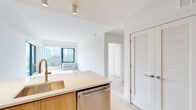 Located at 166 Van Reipen Avenue in Jersey City, NJ, this modern 2-bedroom, 1-bathroom residence offers a thoughtfully designed single-floor layout with a clean, contemporary aesthetic throughout. Light-toned hardwood flooring flows seamlessly across all living areas, complementing the bright white walls and creating a cohesive, airy atmosphere.

The combined kitchen and living area forms the heart of the home. The kitchen is outfitted with stainless steel appliances — including a full-size refrigerator, range, and built-in microwave — alongside two-tone cabinetry featuring white upper cabinets and warm wood lower cabinets, with a clean white tile backsplash. The living area is bathed in natural light from expansive floor-to-ceiling windows and a sliding glass door, framing impressive views of the Jersey City and downtown skyline.

Both bedrooms continue the light wood flooring and neutral palette, with large windows offering additional city views and generous natural light. The primary bedroom is a well-proportioned retreat with ample space. A dedicated closet provides convenient storage.

The bathroom features a sleek, modern design with a navy blue floating vanity, illuminated mirror cabinet, white countertop, and warm-toned tile flooring, offering a polished and functional finish. The overall design emphasizes clean lines, natural light, and urban views throughout.