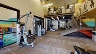 This fitness center offers a comprehensive workout environment spread across a thoughtfully designed multi-level layout. The main floor is generously sized and carpeted with patterned tile flooring, providing a comfortable surface for a wide range of exercises. Cardio equipment, including treadmills and an elliptical trainer by TRUE Fitness, is positioned near large glass partition walls that allow natural light to flow throughout the space. A dedicated water fountain station is conveniently accessible within the cardio area.

The strength training zone is outfitted with cable machines, multi-functional weight stations, and a variety of resistance equipment, offering users a thorough full-body workout experience. A staircase with sleek metal railings leads to an upper mezzanine level, which houses additional weight training equipment and provides an elevated vantage point over the main floor below.

Vibrant, large-scale athletic murals in bold teal, orange, and yellow tones adorn the walls, depicting dynamic silhouettes of athletes in motion, lending an energetic and motivating ambiance to the space. The ceilings feature exposed structural elements on the lower level alongside flush-panel lighting on the upper floor. Windows along the exterior walls bring in glimpses of the surrounding neighborhood, keeping the space feeling open and connected. The overall design balances functionality with an inspiring, active atmosphere.