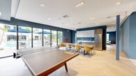 Located at 1162 NE 215th St in Miami, FL, this well-appointed party room offers a thoughtfully designed communal space perfect for hosting events and social gatherings. The interior is defined by a cohesive modern aesthetic, featuring deep navy-blue accent walls, crisp white ceilings with recessed lighting, and light-toned herringbone-patterned flooring that runs throughout the entire space.

The main gathering area includes a dedicated game zone with a full-size ping pong table, complemented by floor-to-ceiling sliding glass doors that open directly to an outdoor pool area, filling the room with abundant natural light and lush tropical views. A dining section features a long communal table with modern amber-hued chairs, ideal for group meals or events.

The kitchenette is equipped with upper and lower cabinetry in a two-tone wood and white finish, a stainless steel refrigerator, and an oven — providing all essentials for catered or self-served gatherings. A separate lounge area offers generous modular seating with teal-trimmed upholstery, framed by large windows overlooking the surrounding greenery. Curated wall art adds a refined touch throughout. The space also includes a bathroom and entrance, completing this versatile and inviting amenity room.