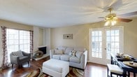 This single-floor space presents a well-appointed living area that balances comfort and functionality. The room is finished with warm hardwood flooring that runs throughout, grounding the space with a classic, inviting feel. Neutral, light beige walls create a bright and airy atmosphere, enhanced by natural light streaming through a set of elegant French doors that open to an outdoor balcony, offering a seamless connection between interior and exterior.

A ceiling fan is centrally positioned, providing both practical airflow and a subtle decorative touch. The living area features a fireplace set against one wall, accompanied by a tall mirror that adds visual depth to the room. Decorative wall art and patterned curtains introduce modest character to the otherwise understated palette.

The layout accommodates comfortable seating, with ample space for a sofa and ottoman arrangement centered on a patterned area rug. The textured ceiling adds a traditional architectural detail overhead. Overall, the space conveys a warm, livable ambiance well-suited for everyday relaxation and entertaining.
