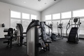 This fitness center offers a generously sized, single-floor layout designed to accommodate a comprehensive range of workout activities. The space is defined by its striking vaulted ceiling, which rises dramatically to create an open, airy atmosphere and contributes to a sense of spaciousness throughout. A ceiling fan is centrally mounted, supporting air circulation during workouts.

Large, multi-pane windows line the perimeter walls, flooding the interior with abundant natural light while providing views of the surrounding exterior. The neutral white walls and light-toned ceiling complement the dark rubberized flooring, which provides both durability and traction underfoot.

The cardio zone features a treadmill, elliptical trainers, a stair climber, and a stationary bike, accompanied by a wall-mounted television for entertainment. The strength training area is equipped with multiple selectorized cable machines and a weight bench, while a dedicated free weights section includes a full dumbbell rack, exercise balls in multiple sizes, and a large floor-to-ceiling mirror that visually expands the space and supports form monitoring.

Recessed lighting supplements natural light for evening or low-light use. The overall design is clean, functional, and well-organized, creating an inviting environment suited to a variety of fitness routines.