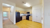 This single-floor residential unit offers a well-organized and functional layout across several distinct living areas. The kitchen is outfitted with light maple cabinetry providing ample storage, dark laminate countertops, and a full suite of black appliances including a refrigerator, electric range, built-in microwave, and dishwasher. Light hardwood-style flooring runs throughout the kitchen and entry areas, creating a clean and cohesive look.

The entry area welcomes residents with hardwood-style flooring, a double closet for storage, and a large window that lets in generous natural light with views of an outdoor balcony and greenery beyond. The living area transitions to plush carpeting underfoot and features a ceiling fan with a light fixture overhead, contributing to a comfortable and airy atmosphere.

The bedroom areas are finished with neutral carpet and clean white walls, offering versatile spaces ready for personalization. Storage is well-addressed throughout, with closet access visible in multiple rooms.

The bathroom includes a wide soaking tub with tiled surround, providing a relaxing retreat. A second bathroom features hardwood-style flooring, a vanity with a dark countertop, an integrated sink, and a large mirror. The overall interior maintains a neutral, light color palette with white trim and doors throughout, lending the space a bright and open feel.
