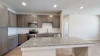 Located at 13003 FM-1488 in Magnolia, Texas, this single-story unit presents a thoughtfully designed, contemporary interior with consistent finishes throughout. The kitchen serves as a standout centerpiece, showcasing rich dark taupe cabinetry extending to the ceiling for generous storage, granite countertops with subtle veining, a classic white subway tile backsplash, and a full suite of stainless steel appliances. A large kitchen island provides additional prep and dining space, while light wood-look flooring anchors the open layout and flows seamlessly into the adjacent living area.

The living space is bright and open, benefiting from multiple windows fitted with blinds that allow natural light to fill the room. A ceiling fan adds comfort, and a door leads directly to an exterior access point. Bedrooms maintain the same light flooring and neutral wall tones, with windows offering outdoor views. A dedicated walk-in closet features multi-tiered wire shelving for organized storage.

The bathroom is well-finished, featuring granite-topped vanity cabinetry, wood-look flooring, white subway tile surrounding a soaking tub and shower combination with brushed nickel fixtures, and direct access to the walk-in closet, creating a practical and cohesive private retreat.