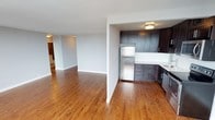 This single-floor residence offers a thoughtfully designed open-concept layout that seamlessly connects the living and kitchen areas, creating a spacious and inviting atmosphere. Rich hardwood flooring flows throughout the unit, adding warmth and a timeless aesthetic to the interior.

The kitchen is a standout feature, equipped with sleek dark-gray cabinetry that extends to the upper and lower walls, providing ample storage space. A classic white subway tile backsplash contrasts beautifully with the dark cabinetry, while stainless steel appliances — including a full-size refrigerator and dishwasher — complete the modern finish. The kitchen opens directly to the main living area through a half-wall partition, maintaining both visual connectivity and functional separation.

The open living area is bright and generously proportioned, with clean white walls and smooth ceilings that enhance the sense of airiness. Neutral tones throughout the space lend themselves well to a variety of furnishing styles. A glimpse down the hallway reveals additional rooms beyond, suggesting a well-organized and practical floor plan. Flush-mount ceiling lighting provides consistent illumination, and the overall design reflects a clean, contemporary residential aesthetic.