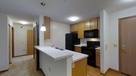 This single-floor space centers around a well-appointed kitchen designed for both functionality and everyday convenience. The kitchen features warm oak cabinetry that lines the upper and lower walls, providing ample storage throughout. Black appliances, including a full-size refrigerator, electric range, and over-the-range microwave, lend a cohesive, classic look to the space.

A peninsula with a white countertop extends outward, creating a natural divider between the kitchen and the adjacent living area. This peninsula also houses a double-basin sink and offers seating potential, making it an ideal spot for casual dining or meal prep. A pendant light fixture hangs above the peninsula, providing focused task lighting that complements the flush-mount ceiling light overhead.

The kitchen flows into a carpeted living area, while a hallway visible in the background suggests additional rooms beyond. The textured ceiling and neutral wall tones contribute to a clean, understated aesthetic throughout. Overall, the space is practical and well-organized, with a layout that maximizes utility while maintaining an open, connected feel between the kitchen and living areas.