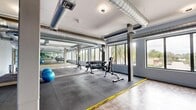 Located at 6250 Griffin Rd in Davie, FL, this fitness center is a well-appointed, single-floor facility designed to support a variety of workout routines. The space is divided into two functional zones: a weight training area and an open exercise floor, clearly delineated by yellow safety markings on the floor.

The weight training area features durable rubber flooring, a dumbbell rack, adjustable weight benches, and exercise balls, providing everything needed for strength and functional training. Large windows along the exterior wall flood this zone with natural light and offer views of the surrounding palm-lined landscape, creating an energizing workout environment.

The open exercise floor transitions to smooth, wood-look vinyl flooring, ideal for stretching, yoga, or group fitness activities. Full-length mirrors line the walls throughout the space, enhancing the sense of openness and allowing users to monitor their form during workouts.

Architecturally, the space features an industrial aesthetic with exposed silver HVAC ductwork running along the white ceiling, supported by structural columns painted in a cool gray-blue tone that complements the facility's overall color palette. Recessed and flush-mounted ceiling lights provide consistent, even illumination throughout. A glass-paneled entry door adds a clean, professional touch to the interior. The overall ambiance is functional, bright, and modern.