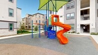 Located at 39261 Delhaven Street in Murrieta, California, this outdoor playground area serves as a well-appointed recreational amenity within a modern multi-story residential community. The space is defined by a vibrant, multi-component play structure finished in bold orange, blue, and green, featuring a spiraling slide, climbing ladders, a lookout tower, and a horizontal monkey bar extension — all sheltered beneath large lime-green shade canopies that provide protection from the Southern California sun. A dedicated swing set with blue posts and toddler-friendly seats complements the main structure, offering age-appropriate play options. The entire play zone is surfaced with rubberized, impact-absorbing flooring in a neutral tone, ensuring both safety and durability. The playground is framed by concrete walkways, low-water landscaping with native grasses and young trees, and stone-accented building facades that reflect the community's contemporary architectural style. Covered parking structures and well-lit pathways are visible in the surrounding area, adding to the functionality of the community. The open-air setting benefits from ample natural light, and the thoughtfully designed layout makes this playground a central and accessible outdoor gathering point for residents.