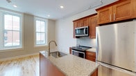 This single-floor space showcases a well-appointed kitchen as its centerpiece. The kitchen is designed with warm medium-toned wood cabinetry that provides ample storage, complemented by granite countertops featuring subtle earth-tone variations. A central island with an integrated stainless steel sink and a sleek pull-down faucet serves as both a functional workspace and a visual anchor for the room.

The cooking area is equipped with a full suite of stainless steel appliances, including an electric range, an over-the-range microwave, and a full-size refrigerator. A mosaic tile backsplash in neutral tones adds texture and visual interest behind the range, tying together the overall design aesthetic.

Natural light filters into the space through a double-hung window, brightening the neutral wall tones and light hardwood flooring that extend throughout. Recessed lighting and a ceiling-mounted track lighting system provide additional illumination, ensuring the kitchen remains well-lit at all times. The overall ambiance is clean, modern, and functional, with quality finishes throughout.