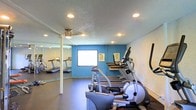 This single-floor fitness space is purpose-built to provide a functional and focused exercise environment. The room features a selection of cardio equipment, including treadmills, an elliptical trainer, and a stationary bike, arranged to maximize use of the available floor space. A full-length mirror along one wall visually expands the room while allowing users to monitor their form during workouts. The bold blue accent wall adds a energizing pop of color to the interior, complementing the neutral tones of the flooring and surrounding walls. Recessed lighting is evenly distributed across the textured ceiling, ensuring the space is well-lit throughout. A small window on the accent wall provides a degree of natural light and ventilation. Motivational posters are displayed on the walls, contributing to an encouraging atmosphere. The durable dark flooring is practical and easy to maintain, well-suited for high-traffic fitness use. Overall, the space is thoughtfully arranged to deliver a straightforward and efficient workout facility.
