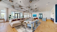 Located at 2420 Medicine Lake Dr in Apopka, Florida, this generously proportioned single-floor clubhouse showcases a sophisticated coastal-modern aesthetic that blends elegance with comfort. The space opens through black-framed glass entry doors into a wide, welcoming corridor defined by bold navy blue shiplap accent walls, warm light wood flooring laid in a distinctive herringbone pattern, and a dramatic cascading chandelier overhead. Large floor-to-ceiling windows line the walls, flooding the interior with natural light and offering views of the surrounding outdoor landscape. The expansive main living area is a standout, featuring an ensemble of sculptural vertigo-style pendant lights suspended from soaring ceilings, creating an artistic focal point above generous seating arrangements that include plush sectional sofas and accent chairs. A large wall-mounted display screen anchors one seating vignette, while a billiards table adds a recreational element to the open floor plan. Curated coastal artwork, open shelving with decorative objects, and a built-in beverage station add refined detail throughout. Separate lounge zones with glass-partitioned walls allow for flexible, multi-purpose use. The overall ambiance is bright, airy, and polished — ideal for community gathering and relaxation.