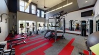 This fitness center is designed to accommodate a wide range of workout routines within a spacious, single-level layout. The room's most striking architectural feature is its double-height ceiling, which creates an open, airy atmosphere enhanced by large windows that line the walls, allowing generous natural light and outdoor views to fill the space. Ceiling fans and industrial-style pendant lighting complement the natural illumination throughout.

The flooring consists of a distinctive patterned carpet in bold red and grey tones, adding visual energy to the space. Along the windowed wall, a row of cardio machines — including treadmills, ellipticals, and stationary bikes — are equipped with individual monitor screens. A large wall-mounted mirror runs along one side of the room, visually expanding the space and providing workout feedback.

The strength and functional training area includes a multi-use cable machine, a mini trampoline, kettlebells, a weight bench, and a wall-mounted horizontal ladder with TRX suspension straps. Stability balls and additional free weights are also available. A full-length mirror panel reflects the free weights section, further enhancing the sense of space. The gym is accessible via a glass-paneled door, reinforcing its open, welcoming character.