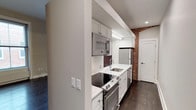 This single-floor space artfully combines contemporary design with timeless character. The kitchen is a standout feature, showcasing white shaker-style cabinetry, crisp white quartz countertops, and a classic subway tile backsplash. Stainless steel appliances — including a built-in microwave, a smooth-top electric range with oven, a refrigerator, and a dishwasher — provide a full suite of modern conveniences. A stainless steel undermount sink adds a clean, polished touch to the workspace. A striking exposed brick column anchors the kitchen, offering a warm, industrial contrast to the otherwise bright and modern palette. Rich, dark hardwood flooring runs throughout, grounding the space with depth and warmth. Recessed lighting ensures the kitchen is well-lit and functional, while the layout is efficiently designed to maximize workspace and flow. The overall ambiance strikes a balance between urban character and contemporary refinement, making the space both highly functional and visually appealing. The neutral white walls and clean architectural lines further enhance the sense of openness and brightness throughout.