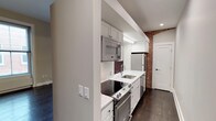 This single-floor space artfully combines contemporary design with timeless character. The kitchen is a standout feature, showcasing white shaker-style cabinetry, crisp white quartz countertops, and a classic subway tile backsplash. Stainless steel appliances — including a built-in microwave, a smooth-top electric range with oven, a refrigerator, and a dishwasher — provide a full suite of modern conveniences. A stainless steel undermount sink adds a clean, polished touch to the workspace. A striking exposed brick column anchors the kitchen, offering a warm, industrial contrast to the otherwise bright and modern palette. Rich, dark hardwood flooring runs throughout, grounding the space with depth and warmth. Recessed lighting ensures the kitchen is well-lit and functional, while the layout is efficiently designed to maximize workspace and flow. The overall ambiance strikes a balance between urban character and contemporary refinement, making the space both highly functional and visually appealing. The neutral white walls and clean architectural lines further enhance the sense of openness and brightness throughout.