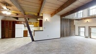 This two-story space presents a compelling mix of rustic and structural design elements throughout. The main level is anchored by a generously proportioned open living area finished with wall-to-wall carpet and a striking reclaimed wood plank ceiling supported by heavy, dark-stained timber beams. Exposed brick columns rise from floor to ceiling, adding raw industrial character and defining the space without fully enclosing it. A chandelier-style light fixture adds a decorative touch overhead. An arched window near the staircase draws in natural light, while glass door panels offer views to the outdoors. A kitchen area is visible beyond the brick columns, featuring dark wood cabinetry.

An open iron-railed staircase with twisted balusters ascends to the second floor, where a landing overlooks the main level. The upper floor features carpeted hallways, wood-paneled walls, and ceiling fans throughout. Two bathrooms are present in the space: one featuring tile flooring, a tub-shower combination, and a wood-framed vanity, and another with large-format tile floors, pendant lighting, and a wide wood vanity. Together, the two levels offer a layered, characterful environment with notable architectural detail.