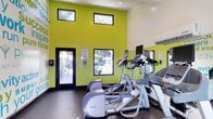 This fitness center is designed to energize and motivate with its bold lime-green accent walls adorned with inspirational text and graphics. The space benefits from soaring ceilings that create an open, airy atmosphere, while a row of clerestory windows near the top of the walls invites abundant natural light without sacrificing wall space for equipment. Additional windows at a lower level offer views of an outdoor pool area, seamlessly connecting the interior to the surrounding landscape. The facility is outfitted with a selection of professional-grade cardio equipment, including Precor elliptical trainers and treadmills, each equipped with integrated display screens for an engaging workout experience. A large wall-mounted mirror expands the sense of space and allows users to monitor their form. The dark, speckled flooring provides a durable, easy-to-maintain surface suited for high-traffic use. A keycard-access entry door ensures secure, controlled access for authorized users. The overall design balances functionality with an energetic aesthetic, making it a practical and visually stimulating environment for fitness activities.