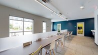 This single-floor space is designed as a functional and contemporary shared workspace. The open-plan layout accommodates multiple long workstation tables arranged in parallel rows, each paired with modern Eames-style chairs in white and subtle yellow accents, creating a clean and inviting atmosphere.

The interior design balances neutral tones with a bold teal accent wall that anchors one end of the room, adding visual depth and energy to the space. The opposite wall features large windows that allow generous natural light to fill the room, enhancing the bright, airy feel. The white ceiling is fitted with sleek linear LED lighting strips and track lighting, ensuring consistent illumination throughout.

A low partition divider separates the main workspace from a smaller, semi-private seating area toward the rear, offering flexibility for group collaboration or focused individual work. The flooring consists of neutral-toned carpet tiles, contributing to a quiet, professional environment. Built-in power access points are integrated into the workstation surfaces, supporting productivity needs. Overall, the space strikes a balance between modern aesthetics and practical functionality.