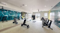 Located at 9015 Eager Rd in Richmond Heights, Missouri, this well-appointed fitness center offers a comprehensive workout environment across a single open floor. The space is thoughtfully designed with distinct functional zones that cater to a variety of exercise routines. A bold teal accent wall emblazoned with the motivational phrase "Don't Stop. Turn It Up!" anchors the room, setting an energetic tone throughout. A row of spin bikes lines the wall adjacent to this graphic mural, creating a dedicated cycling area. Free weights, including a tiered dumbbell rack, and an assortment of stability balls provide options for strength and functional training. Large floor-to-ceiling windows along the exterior walls flood the space with natural light and offer views of an outdoor courtyard and pool area. Full-length mirrors on one wall enhance the sense of openness and allow for form-checking during workouts. A ceiling fan maintains comfortable airflow throughout the room. A separate studio area, accessed through glass-panel doors, features a patterned accent wall, spin bikes, yoga mats, and foam blocks, making it ideal for stretching, cycling, or group fitness sessions. Recessed lighting and a clean, neutral color palette complete the modern, polished aesthetic of this fitness facility.