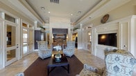 This single-floor community amenity space showcases a refined, traditional design aesthetic defined by travertine tile flooring, tray ceilings with recessed lighting, and crisp white millwork and crown molding throughout. The central lounge area serves as the heart of the space, anchored by a striking stacked-stone fireplace surround that extends to the ceiling, flanked by patterned wingback chairs and a dark wood coffee table atop a woven area rug. A wall-mounted flat-screen television is framed by detailed wainscoting panels, adding a polished focal point to the seating area.

Glass-paned French doors connect the various zones of the space, maintaining visual openness while defining distinct areas. A dedicated game room features a full-sized billiards table illuminated by pendant drum lights, with natural light streaming through roman shade-dressed windows. A shuffleboard table is also present, enhancing the recreational offerings.

A café-style seating area with bistro-height tables and upholstered chairs lines a bank of large windows that overlook an outdoor pool and landscaped grounds, filling the space with abundant natural light. A kitchenette or bar area with dark cabinetry is visible in the background, complementing the overall warm, neutral color palette that gives the entire space an inviting, upscale ambiance.