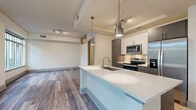 Located at 2121 North Westmoreland Street in Arlington, VA, this thoughtfully designed one-bedroom, one-bathroom residence offers a modern aesthetic with high-quality finishes throughout. The open-concept layout seamlessly connects the kitchen and living area, creating an inviting and functional space. The kitchen is a standout feature, boasting a large quartz waterfall island with an undermount sink, two-tone cabinetry in white and dark gray, stainless steel appliances including a range, microwave, refrigerator, and dishwasher, plus a classic subway tile backsplash. A tray ceiling with track lighting and elegant pendant lights adds architectural interest overhead. Hardwood-style flooring flows throughout the main living areas, complementing the clean, contemporary palette. The bedroom is a comfortable retreat featuring a ceiling fan, a transom window for additional natural light, and direct access to a spacious walk-in closet with wire shelving. The bathroom is finished with large-format tile flooring, dark gray cabinetry, and a well-lit vanity mirror. A dedicated laundry area houses a full-size washer and dryer with built-in shelving. A hallway lined with natural light leads to views of lush greenery, adding a serene outdoor connection to this well-appointed urban residence.