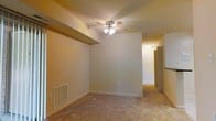 This single-floor space presents a straightforward, functional layout with warm, neutral-toned walls throughout. The main living area is finished with wall-to-wall carpet in a light beige tone, providing a soft underfoot feel. A ceiling fan with an integrated light fixture serves as the central overhead lighting source, offering both illumination and air circulation. A floor-level HVAC vent is positioned along the base of one wall, ensuring climate control throughout the space. The walls are kept clean and simple, with standard white baseboards framing the room. A structural soffit detail is visible near the ceiling in one corner, adding a subtle architectural element to the otherwise open room. A hallway extends from the main living area, leading to additional rooms and providing a sense of depth to the layout. The corridor is illuminated by a flush-mounted ceiling light, keeping the passage well-lit. Neutral, consistent finishes are carried throughout, creating a cohesive and understated aesthetic. The overall ambiance is modest and practical, suited for comfortable everyday living.