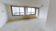 This single-floor space features a generously sized open living area finished with plush, wall-to-wall carpeting in a neutral cream tone. The room is defined by a wide bank of floor-to-near-ceiling windows with dark bronze-toned frames, which span much of the exterior wall and flood the interior with abundant natural light. The windows offer elevated views of the surrounding urban skyline, with neighboring high-rise buildings visible in the backdrop. A sliding glass door positioned to one side of the window bank provides direct access to a balcony, further enhancing the connection to the outdoors and extending the sense of openness. A baseboard heating unit runs beneath the windows, offering a practical climate control solution integrated seamlessly into the room's layout. The walls and ceiling are finished in a clean, neutral white, contributing to a bright and airy atmosphere throughout. A ceiling-mounted light fixture provides overhead illumination, complementing the natural light. The overall ambiance is calm and understated, presenting a versatile, move-in-ready canvas well-suited for a variety of furnishing styles and layouts.