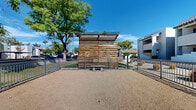 Located at 8560 W Peoria Ave in Peoria, Arizona, this outdoor dog park is a dedicated amenity space designed to provide a safe and functional environment for pets and their owners. The single-level area is fully enclosed by metal rail fencing, offering secure boundaries on all sides with gated access points for easy entry and exit. The ground is covered with a natural gravel and compacted dirt surface well-suited for the arid Arizona climate, requiring minimal maintenance while remaining practical for active use. A standout architectural feature is the slatted wooden pergola structure positioned at one end of the park, constructed with horizontal wooden planks and a latticed overhead canopy that provides partial shade and a visual focal point. Concrete benches are placed within the enclosure, offering a comfortable resting spot for owners while their pets roam freely. A pet waste station, complete with a dispenser and waste receptacle, is conveniently located near the entry area, promoting cleanliness and responsible pet ownership. Mature trees surrounding the perimeter provide natural shade and a pleasant green backdrop against the bright Arizona sky. The park sits adjacent to a multi-story residential complex with a Southwestern-inspired stucco exterior, integrating seamlessly into the broader community landscape.