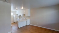 This single-floor space features an open-concept layout that seamlessly connects the kitchen and living area, creating a practical and cohesive environment. The kitchen is outfitted with white cabinetry that extends to the ceiling, providing ample storage, while a peninsula counter offers additional workspace and serves as a natural divider between the kitchen and the adjacent living area. The kitchen includes a white refrigerator, a gas range, and a stainless steel sink, all arranged efficiently within a compact footprint.

Warm hardwood-style flooring runs throughout the space, lending a sense of continuity and warmth to the interior. The neutral beige walls complement the light-toned surfaces and white finishes, contributing to a clean, understated aesthetic. Ceiling-mounted light fixtures provide consistent overhead illumination throughout both the kitchen and living areas.

The living area is open and unobstructed, offering flexibility for furniture arrangement. White baseboards frame the perimeter of the room, adding a crisp finishing detail. Overall, the space presents a straightforward, functional design well-suited for everyday living.