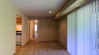 This single-floor residential space offers a straightforward and functional layout designed for comfortable everyday living. The main living area features warm-toned carpeting underfoot and neutral taupe walls that create a subdued, inviting atmosphere. A structural beam detail is visible along the ceiling, adding a subtle architectural element to the room. Recessed lighting provides focused illumination from above, complementing the natural light that filters in through a large window dressed with vertical blinds along one wall.

A baseboard heating unit is positioned beneath the window area, indicating a practical heating setup throughout the unit. The flooring transitions to carpet as the space continues down a hallway, which leads to additional rooms visible in the background. The hallway connects to what appears to be a bedroom with a window allowing natural light into the corridor.

The overall aesthetic is clean and minimal, with white trim and baseboards providing a crisp contrast against the neutral wall tones. The space presents an open, uncluttered feel with ample wall space and functional built-in features. The single-floor layout ensures seamless accessibility throughout, making it a practical and livable environment suited for a range of occupants.