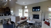 This well-appointed single-floor residence showcases a thoughtfully designed living space that blends modern aesthetics with comfortable functionality. The living room serves as the centerpiece of the home, boasting elevated ceilings accented by crisp crown molding that adds a refined architectural touch. Large windows allow generous natural light to fill the room, framed by patterned drapes that introduce a playful design element against the neutral, warm-gray walls. A sleek, tile-surround fireplace anchors one wall and is complemented by a wall-mounted flat-screen television above, creating a natural focal point for the room. The space is furnished with a clean-lined white sofa adorned with rust-toned accent pillows, paired with a round white coffee table that sits atop a striking black-and-white geometric area rug, lending a bold contemporary flair to the interior. Recessed lighting in the ceiling supplements the abundant natural light, ensuring the space is bright and inviting at any time of day. The overall ambiance is modern yet warm, with a careful balance of neutral tones and vivid accents that give the home a polished, curated character.