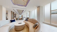Located at 2100 N Central Rd in Fort Lee, NJ, this generously sized single-floor amenity space offers a sophisticated, modern environment designed for both relaxation and productivity. The open layout is anchored by a sweeping lounge area featuring curved sectional sofas, rounded accent tables with wood-paneled bases, and layered ambient lighting including dramatic circular pendant fixtures and purple-accented cove lighting set within tiered ceiling coffers. Floor-to-ceiling windows line the perimeter, flooding the interiors with abundant natural light and views of the surrounding greenery. The living and lounge areas transition seamlessly into a formal dining space furnished with varied seating arrangements atop textured carpeting beneath an industrial-chic circular chandelier and a polished concrete ceiling accent. Glass-walled conference rooms provide private meeting spaces while maintaining visual connectivity to the open areas, complemented by curated artwork displayed throughout the corridors. The kitchen features sleek cabinetry, a white quartz countertop, and bar seating with woven leather chairs. The game room stands out with light hardwood flooring, a golf simulator, a foosball table, and a billiards table, all surrounded by comfortable lounge seating and bright natural light — making it an exceptional entertainment destination.