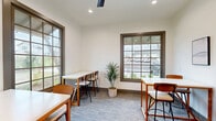 Located at 988 Old Shirley Rd in Central, SC, this single-room study and co-working space offers a well-appointed environment designed for productivity and collaboration. The room is finished with crisp white walls and neutral-toned carpeting, creating a bright and clean atmosphere throughout. Multiple white-topped high tables paired with warm wood-framed chairs are thoughtfully arranged to accommodate both individual work and small group sessions, providing flexible seating options across the space. Large grid-paned windows with dark frames line two walls, flooding the interior with generous natural light and offering views of the surrounding exterior. Recessed ceiling lighting and a ceiling fan ensure the space remains well-lit and comfortable at all times. A decorative potted palm plant adds a touch of greenery, complementing the minimalist aesthetic. On one wall, a trio of framed abstract artworks contributes a subtle artistic element to the otherwise functional setting. A doorway connects the room to an adjacent area with hardwood flooring, suggesting it is part of a larger facility. The overall ambiance is modern, clean, and purposefully designed to support focused work in a welcoming community environment.