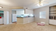 This single-floor residence features an open-concept layout that seamlessly connects the living area and kitchen, creating a spacious and functional flow. Crown molding runs throughout the main living space, adding a refined architectural detail to the neutral-toned walls and light beige carpeting underfoot.

The kitchen is well-appointed with white raised-panel cabinetry extending to the ceiling, offering generous storage capacity. Dark granite-style countertops provide a contrasting visual element, while tile flooring, a white electric range, and a stainless steel sink complete the functional workspace. A peninsula counter separates the kitchen from the living area, serving as both a practical workspace and a casual dining surface.

The living area is bright and open, benefiting from natural light through a window with blinds. A ceiling fan with a light fixture provides both airflow and overhead illumination. Built-in shelving is integrated into one wall, offering dedicated display and storage space. A pendant light fixture and an entry door with secure hardware are visible from the dining area, which also provides access to what appears to be a walk-in closet or storage room with built-in shelving.

The bathroom features tile flooring, a full tub-shower combination, a toilet, and a vanity, all finished in a clean, neutral palette.