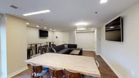 This single-floor communal space is thoughtfully designed to accommodate a variety of social and collaborative activities. The open-plan layout is anchored by a large, light-toned wooden dining table surrounded by warm walnut-finish chairs, making it ideal for group gatherings or shared meals. A plush, dark sectional sofa paired with a rustic coffee table defines a dedicated lounge area, offering a comfortable spot for relaxation or casual conversation. Multiple flat-screen TVs are mounted on the walls throughout the room, ensuring excellent visibility from every seating zone. High-top bar tables and stools are positioned along one wall, providing an additional area for informal seating or socializing. The space is finished with rich hardwood-style flooring that runs throughout, complementing the clean white walls and adding warmth to the overall atmosphere. Lighting is a combination of recessed ceiling fixtures and a central drum pendant light, creating a bright and inviting ambiance. The layout is open and well-organized, with clearly defined functional zones that flow seamlessly into one another, making this a versatile and welcoming space for a range of uses.