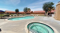 This single-story property showcases a well-appointed outdoor recreational area centered around a generously sized, freeform swimming pool with crystal-clear turquoise water. Adjacent to the pool sits a bubbling hot tub/spa, perfect for relaxation. The expansive concrete sundeck provides ample space for lounge chairs, offering residents and guests comfortable areas to sunbathe and unwind. A shaded patio area with an umbrella-covered table adds a functional gathering spot within the pool enclosure. The community buildings surrounding the pool area are finished in a warm stucco exterior and topped with distinctive red Spanish-style tile roofing, lending the property a Southwest or Mediterranean aesthetic. Mature palm trees and deciduous trees are scattered throughout the grounds, providing natural shade and visual interest against the open blue sky. A white metal perimeter fence encloses the pool area, ensuring privacy and security. A dedicated clubhouse or amenity building with sliding glass doors is accessible directly from the pool deck, further enhancing the functionality of the outdoor space. Overall, the property exudes a relaxed, resort-like ambiance suited for warm-climate living.