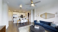 This single-floor residence showcases a thoughtfully designed open-concept layout that seamlessly integrates the kitchen, dining, and living areas into one cohesive, flowing space. The kitchen is outfitted with stainless steel appliances, white cabinetry, a tile backsplash, and a breakfast bar illuminated by elegant pendant lighting, providing both functionality and style. Adjacent to the kitchen, the dining area comfortably accommodates a full-sized table and chairs, making it ideal for entertaining or everyday meals. The living room is anchored by rich, dark hardwood flooring and features a ceiling fan with integrated lighting, wall sconces, and generous wall space accented by a large mirror, contributing to a bright and open ambiance. Neutral-toned walls throughout the space create a clean, modern backdrop that complements the contemporary furnishings. The open flow between all main areas enhances the sense of spaciousness, while the cohesive design language ties the entire floor plan together with a polished, welcoming aesthetic suited to modern living.