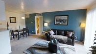 This single-floor residence offers a welcoming and well-designed layout with a distinct sense of comfort and style. The living area serves as the heart of the space, highlighted by a bold teal accent wall that adds depth and character to the room. Plush wall-to-wall carpeting underfoot provides warmth, while a textured area rug anchors the seating arrangement centered around a leather sofa and ottoman. A framed cityscape artwork adorns the accent wall, adding a urban sophistication to the space. A floor lamp provides ambient lighting, complementing the natural light that filters through the room. The textured ceiling adds a classic residential touch overhead. Adjacent to the living area, a dining space is seamlessly integrated, featuring a dark wood table and chairs beneath a pendant light, creating a defined yet open area for meals. A doorway from the living room leads further into the home, hinting at additional rooms beyond. The cohesive color palette of teal, dark brown, and neutral tones throughout the shared living and dining areas creates a harmonious and inviting atmosphere, making this space both functional and visually engaging.