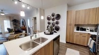 This single-story residence offers two bedrooms and two bathrooms within a thoughtfully designed layout. The kitchen serves as a central highlight of the space, showcasing warm wood-tone cabinetry that extends from lower cabinets to a tall pantry-style storage unit, providing ample storage throughout. Quartz-style countertops in crisp white offer a clean contrast to the natural wood tones, while a deep double-basin stainless steel sink adds functional appeal. Stainless steel appliances, including a dishwasher, complement the modern aesthetic. Pendant lighting hangs above the kitchen island, complementing the track lighting overhead to create a well-lit workspace. The open sightlines from the kitchen connect seamlessly to adjacent living areas, enhancing the sense of flow throughout the home. Wood-look flooring runs through the main areas, adding warmth underfoot. Decorative wall accents, including an arrangement of dark circular art pieces, contribute character to the neutral palette. A glimpse into the adjoining rooms reveals continued modern styling with natural light filtering through, reinforcing the cohesive and contemporary design throughout the residence.