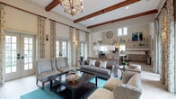 This single-story home showcases a beautifully designed open-concept layout that connects the living area to the kitchen, creating a cohesive and inviting atmosphere. The living room draws immediate attention with its striking exposed wood ceiling beams, which add rustic warmth and architectural character to the otherwise bright, neutral space. An ornate wrought-iron chandelier hangs centrally, complementing the beams and enhancing the room's traditional charm. Tall French doors and large windows dressed in patterned drapery flood the space with natural light, highlighting the teal area rug and carefully curated mix of upholstered seating, including wingback chairs, a tufted sofa, and wicker accent chairs. A dark wood coffee table anchors the seating arrangement. A large decorative clock serves as a focal point along the wall leading into the kitchen, which features white cabinetry, open shelving, and pendant lighting above what appears to be a breakfast bar or island. The overall ambiance is warm yet refined, blending rustic and transitional design elements for a comfortable and stylish living environment.