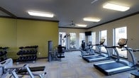 This fitness center offers a comprehensive workout environment designed to support a wide range of exercise routines. The space is illuminated by a combination of large windows that invite natural light and flush-mounted fluorescent ceiling fixtures, creating a bright and energizing atmosphere throughout. A ceiling fan adds to the comfort of the room during intense workouts.

The cardio section is well-stocked with treadmills, an upright stationary bike, and an elliptical machine, all positioned to take advantage of the natural light and outdoor views provided by the generous window placement. A wall-mounted flat-screen television is conveniently situated for entertainment during cardio sessions.

On the strength training side, the space includes a multi-function cable and weight machine, a dedicated weight bench, and a full rack of dumbbells in varying sizes, catering to both beginner and experienced fitness enthusiasts. The flooring consists of durable, patterned carpet tiles suited for a gym environment.

The interior design features warm yellow accent walls contrasted by neutral tones, lending the space a modern and motivating aesthetic. Crown molding adds a refined architectural touch, elevating the overall finish of the room. The layout is thoughtfully organized to allow ample circulation space between equipment.