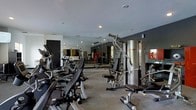 This modern fitness center is thoughtfully designed to support a comprehensive workout experience. The space is outfitted with a generous selection of cardio equipment, including treadmills, ellipticals, and stationary bikes, all arranged for easy access and circulation. On the strength training side, a multi-station cable machine anchors the room alongside various resistance and weight machines suited for a full-body workout. The flooring features durable, cushioned carpet ideal for a gym environment, providing comfort and stability during exercise. Floor-to-ceiling mirrors line one wall, visually expanding the space and allowing users to monitor their form throughout their sessions. The interior is finished with a contemporary palette of dark and neutral tones, giving the space a sleek, professional atmosphere. Recessed lighting illuminates the room evenly, ensuring a bright and functional environment. Motivational artwork adorns the walls, adding a dynamic visual element to the overall design. The layout is efficiently organized to maximize usable floor space while maintaining clear pathways between equipment, making the fitness center both practical and inviting for users of all fitness levels.