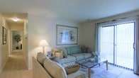 This single-floor space offers a warm and inviting atmosphere characterized by a thoughtfully arranged living area. The living room serves as the centerpiece, furnished with plush, neutral-toned sofas adorned with coordinating accent pillows in soft teal and earth tones. A glass-top coffee table anchors the seating arrangement, adding a touch of elegance to the relaxed setting. A framed piece of abstract artwork decorates the wall above the sofa, contributing a subtle artistic flair to the space. A classic table lamp casts a warm, ambient glow that complements the natural light streaming in through a floor-to-ceiling sliding glass door fitted with vertical blinds, hinting at an outdoor area beyond. The flooring features a light tile pattern that enhances the airy feel of the room. The textured ceiling adds a traditional architectural detail consistent throughout the space. Overall, the space exudes a calm, comfortable ambiance with a neutral color palette that creates a welcoming and livable environment, well-suited for both relaxing and everyday living.