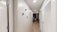 This thoughtfully designed single-floor residence combines contemporary aesthetics with functional living spaces. The layout flows from an entry hallway — accented with decorative wall-mounted hooks and recessed lighting — through to the main living areas. Light wood-tone flooring runs throughout, creating a warm, cohesive aesthetic.

The kitchen is a standout feature, outfitted with gray cabinetry, white subway tile backsplash, and quartz countertops. Stainless steel appliances, including a French door refrigerator, electric range, microwave, and dishwasher, complement the space. A central island with bar seating and a matte black faucet at the sink add both style and practicality. Open shelving provides additional display and storage options, and floor-to-ceiling windows allow city views to serve as a striking backdrop.

The bedroom is a serene retreat, featuring warm neutral tones, a sliding glass barn door, and tasteful décor. It offers ample room for furnishings and connects conveniently to the rest of the unit.

The bathroom is clean and modern, featuring a frameless glass walk-in shower with white tile surround, matte black fixtures, and a wood-toned vanity with a marble-look countertop. The space exudes a spa-like calm. Overall, the residence presents a polished, contemporary living environment.