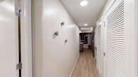 This thoughtfully designed single-floor residence combines contemporary aesthetics with functional living spaces. The layout flows from an entry hallway — accented with decorative wall-mounted hooks and recessed lighting — through to the main living areas. Light wood-tone flooring runs throughout, creating a warm, cohesive aesthetic.

The kitchen is a standout feature, outfitted with gray cabinetry, white subway tile backsplash, and quartz countertops. Stainless steel appliances, including a French door refrigerator, electric range, microwave, and dishwasher, complement the space. A central island with bar seating and a matte black faucet at the sink add both style and practicality. Open shelving provides additional display and storage options, and floor-to-ceiling windows allow city views to serve as a striking backdrop.

The bedroom is a serene retreat, featuring warm neutral tones, a sliding glass barn door, and tasteful décor. It offers ample room for furnishings and connects conveniently to the rest of the unit.

The bathroom is clean and modern, featuring a frameless glass walk-in shower with white tile surround, matte black fixtures, and a wood-toned vanity with a marble-look countertop. The space exudes a spa-like calm. Overall, the residence presents a polished, contemporary living environment.